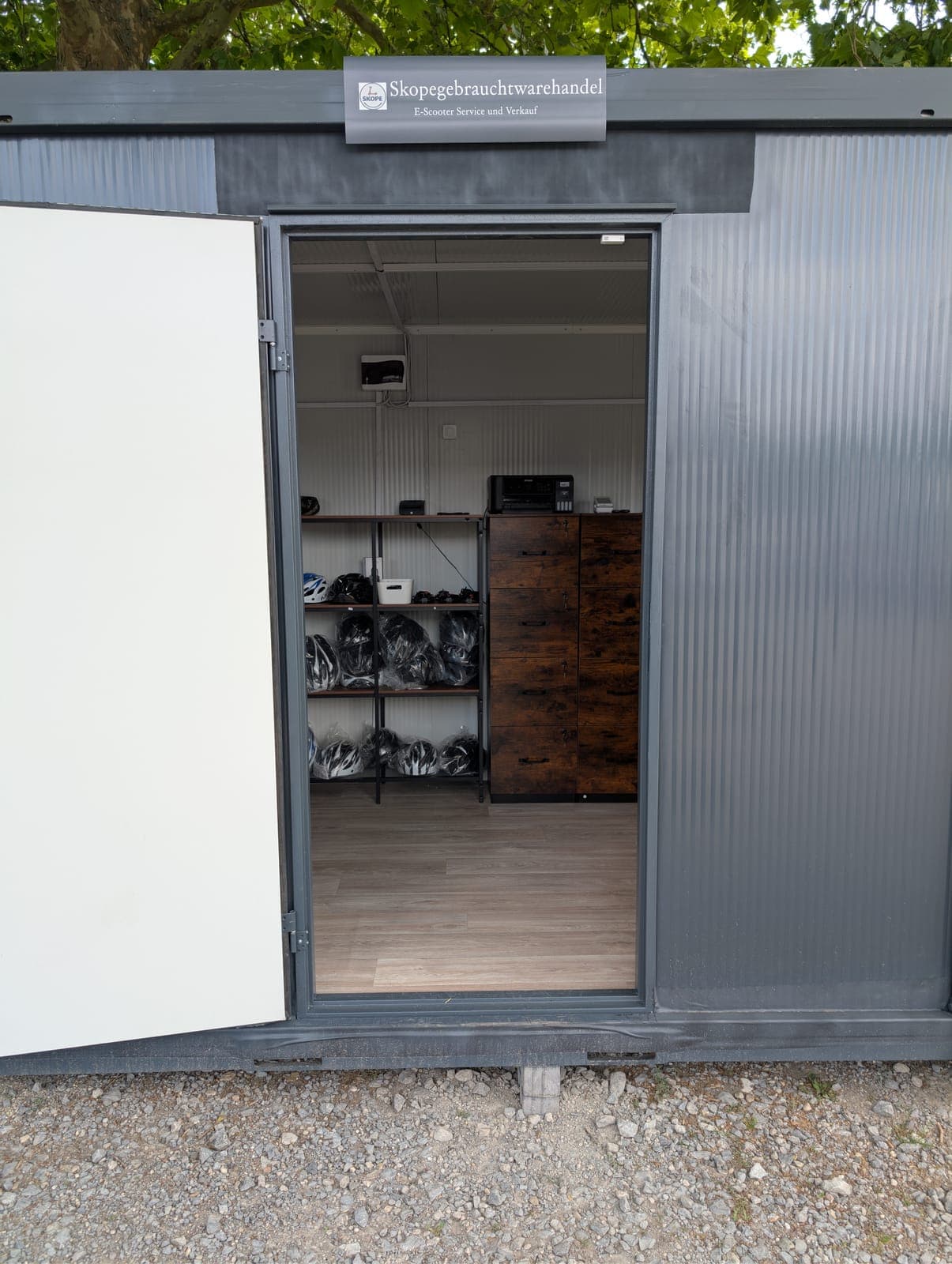 Offene Tür des Bürocontainers unter dem Schild „Skopegebrauchtwarehandel – E-Scooter Service und Verkauf“, innen ein Regal mit verpackten Helmen und ein Aktenschrank