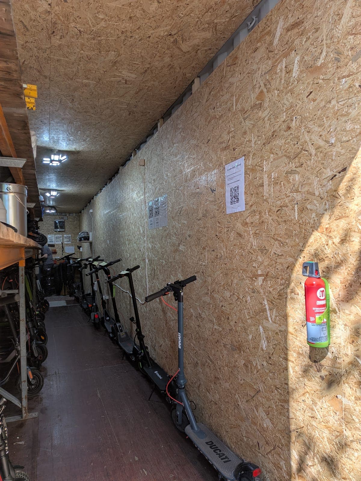 Langer Gang im Lagercontainer mit Wänden aus Grobspanplatten, rechts ein Feuerlöscher, links eine Reihe aufgereihter E-Scooter