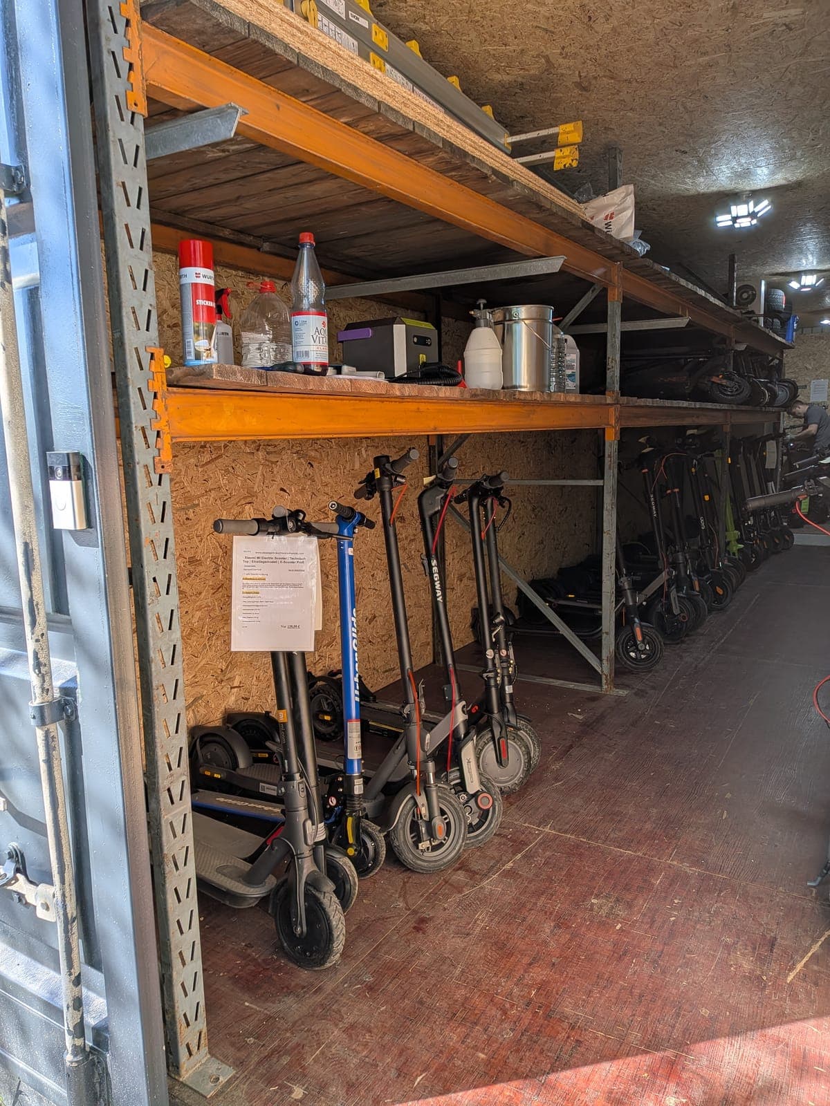 Blick in den geöffneten Lagercontainer: E-Scooter in zwei Reihen unter einem Schwerlastregal, am vordersten Gerät hängt ein bedrucktes Datenblatt am Lenker