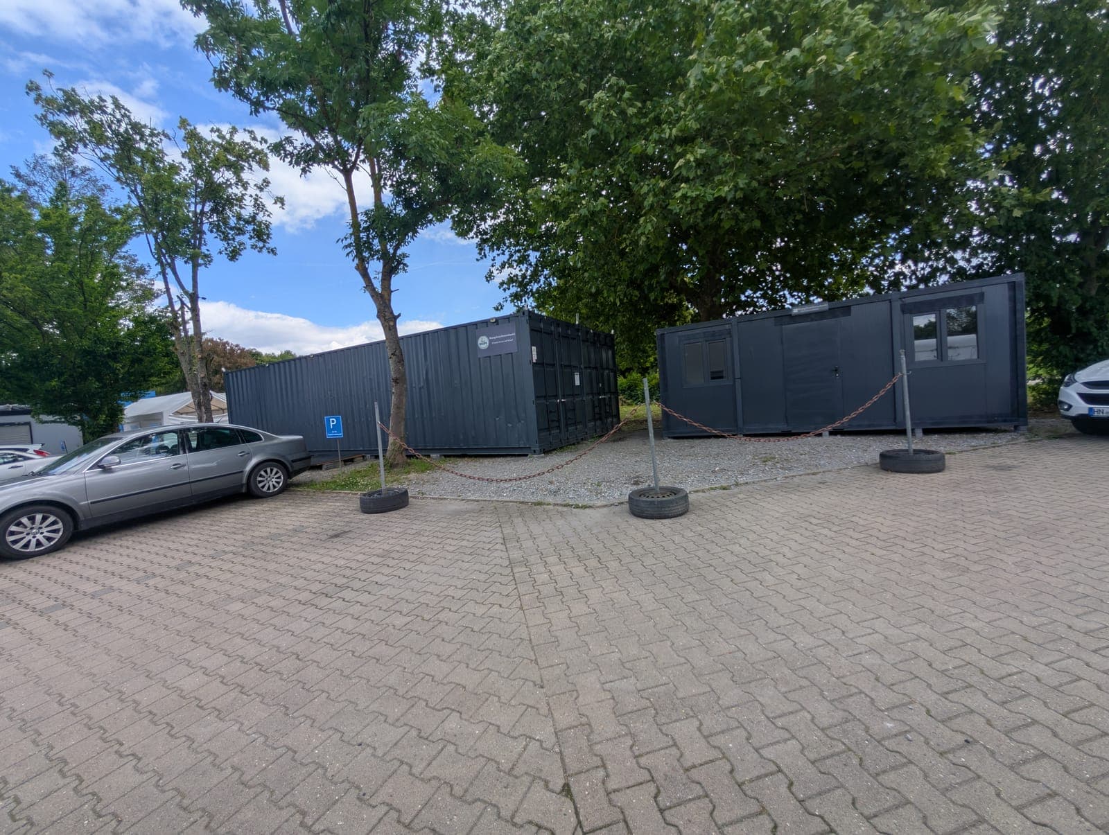 Zwei dunkelgraue Container auf einem gepflasterten Platz unter Bäumen, davor ein mit Ketten abgesperrtes Kiesfeld und Parkplätze
